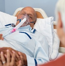 Photograph of a man lying in a bed wearing an oxygen mask.